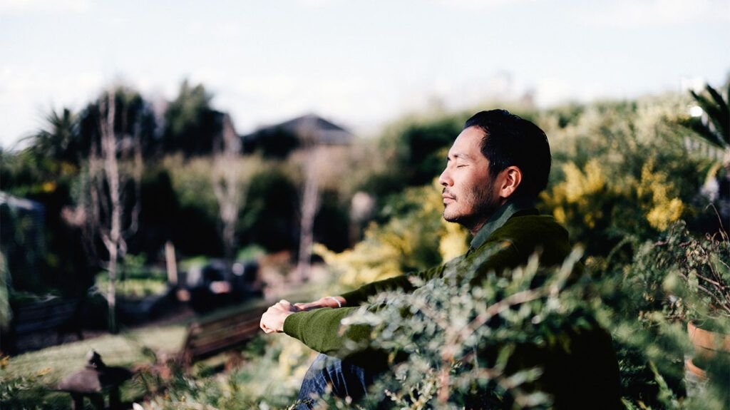 A man sitting in a garden amongst bushes with his eyes closed.